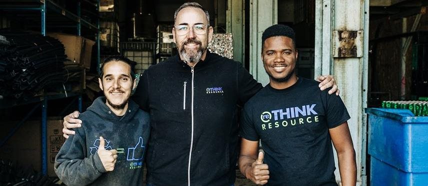 Three people standing together in an industrial workspace wearing RETHINK RESOURCE shirts.