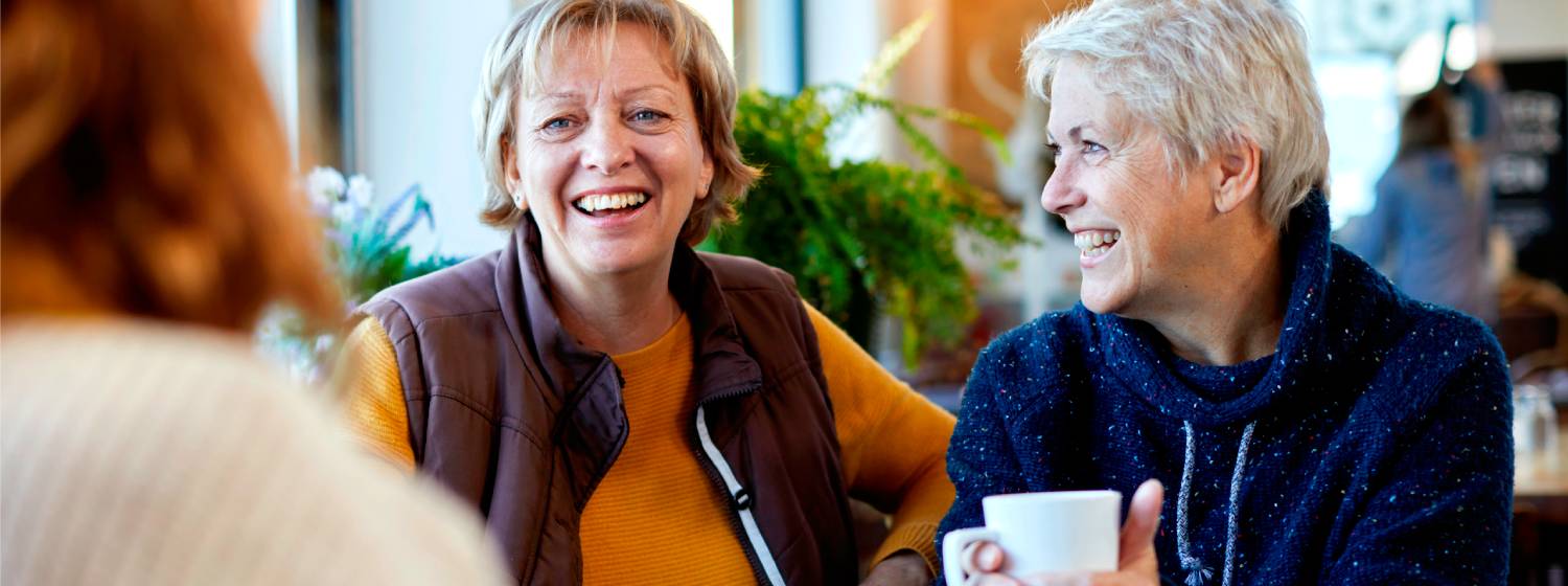 A group of older women sit in a café, smiling and chatting.