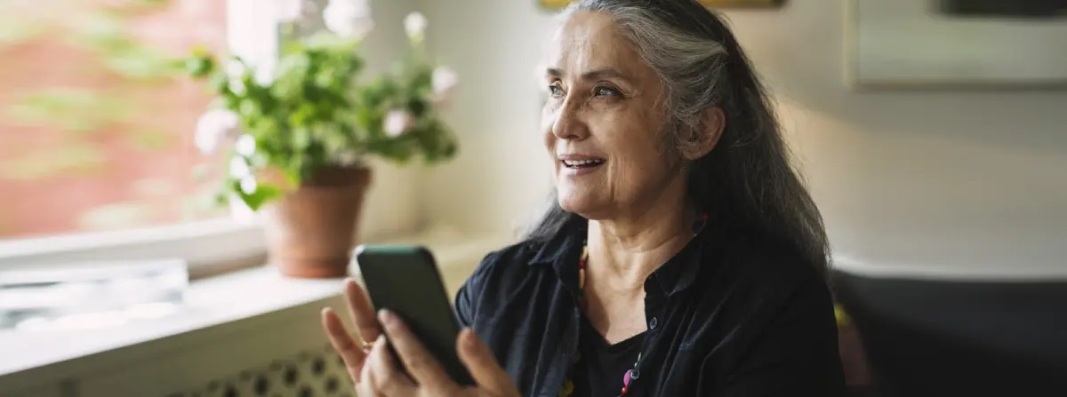 A mature woman with medium skin tone and long, greying hair, holds her phone in front of her, smiling slightly as she speaks. With flowers on the windowsill beside her, she looks out the window