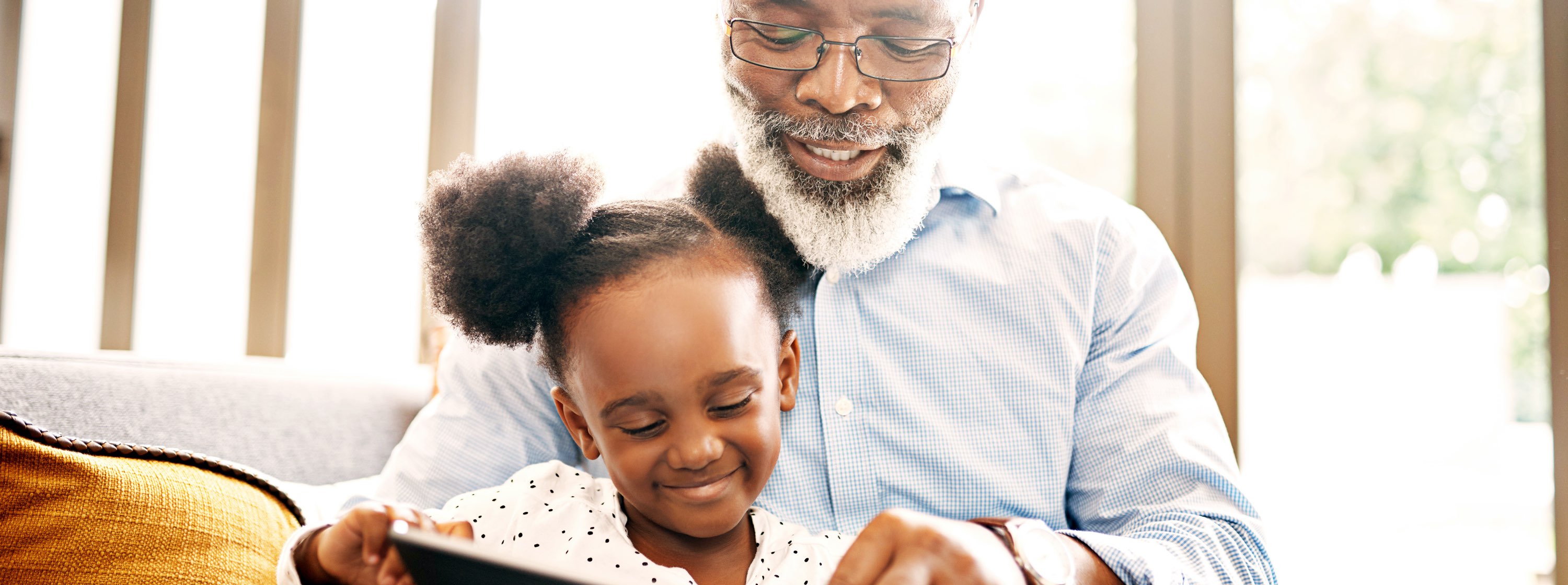A grandfather and his granddaughter sit together, smiling, reading a book.