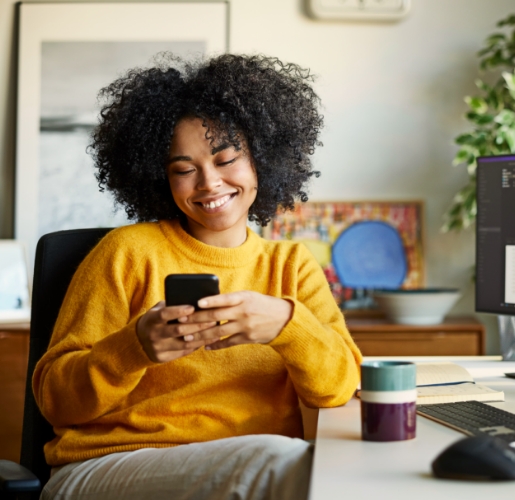 Young black woman sitting at her computer desk and smiling at her phone
