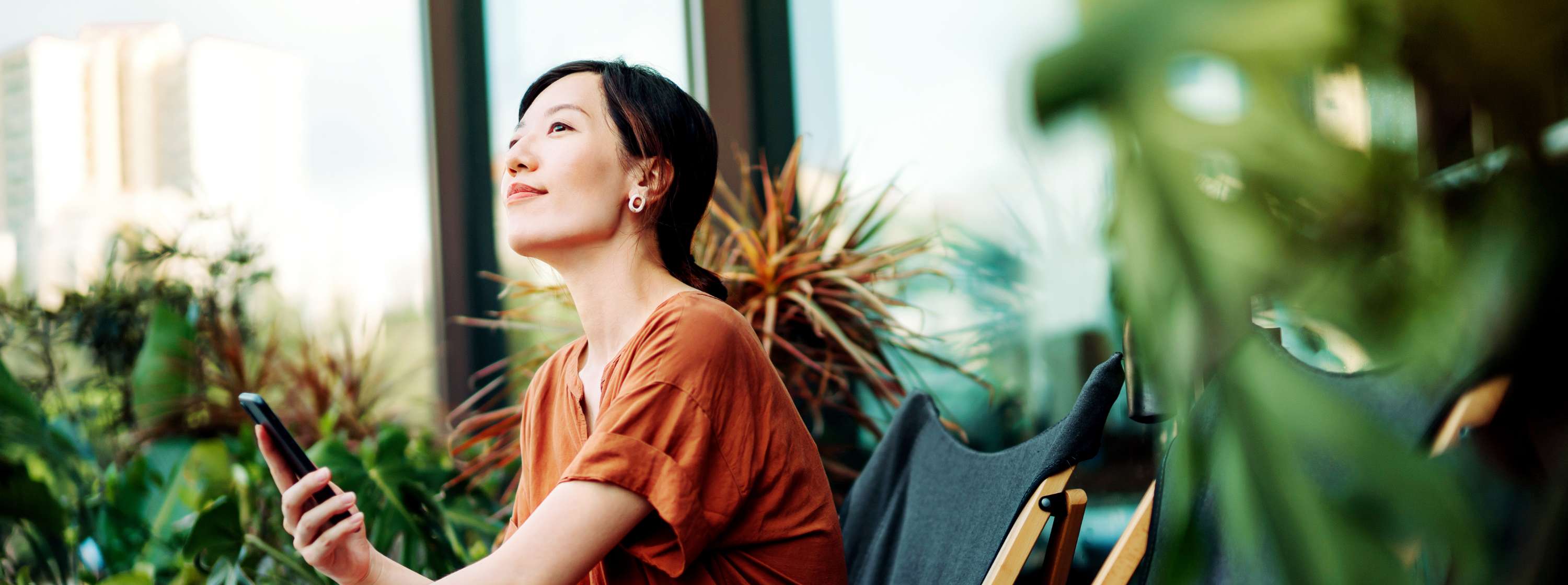 A woman sits in a lush garden. She is holding her mobile phone and looking contemplative.