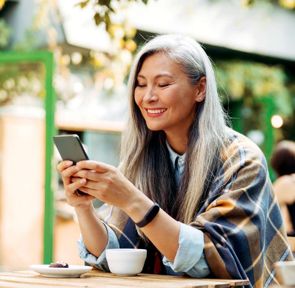 A mature woman with long grey hair sits on a patio at a café, smiling as she looks at her mobile phone.