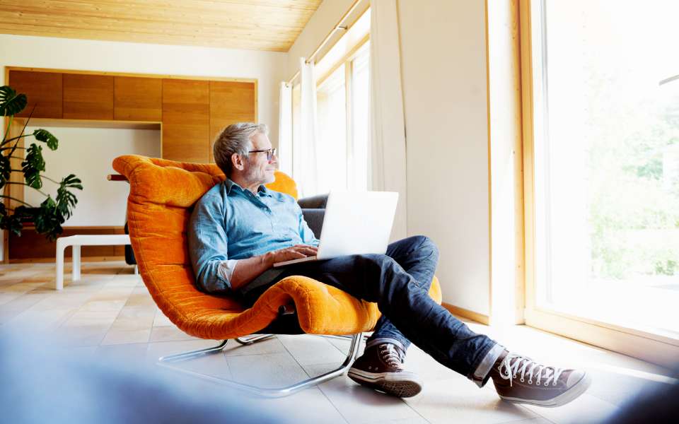 A man lounges in a comfortable chair with his lap top, gazing out the window as light pours in. 