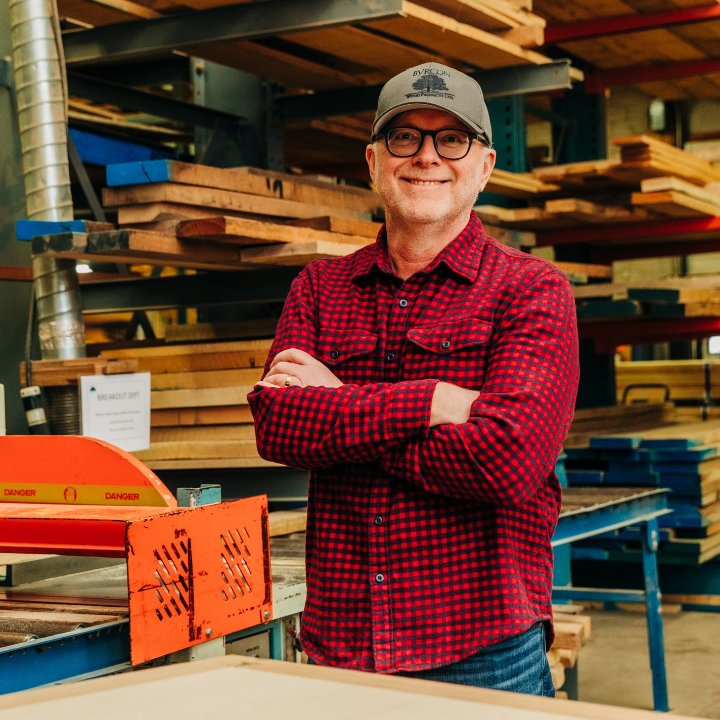 A man wearing glasses, a cap, and a red plaid shirt stands with arms crossed in a woodshop. Byrcon Wood Products. Member since 2014