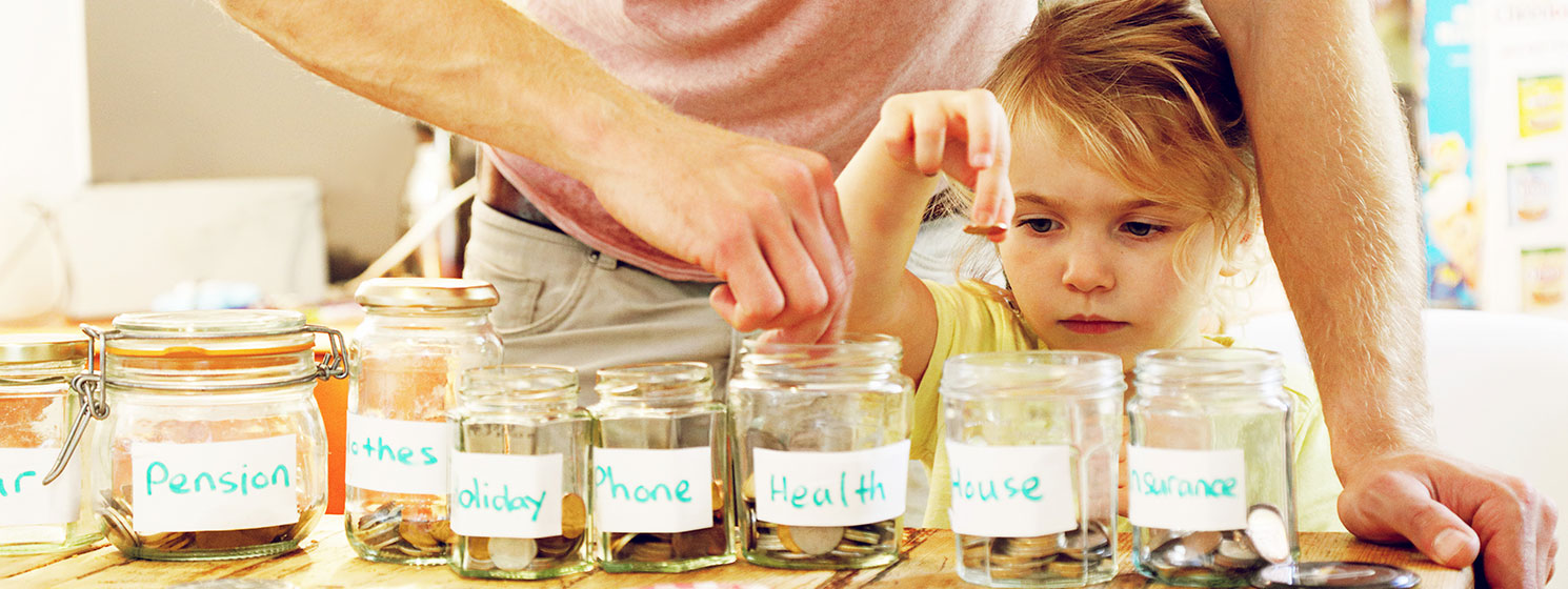 Father teaching daughter how to save using jars marked with different savings goals like pension, holiday, phone and house