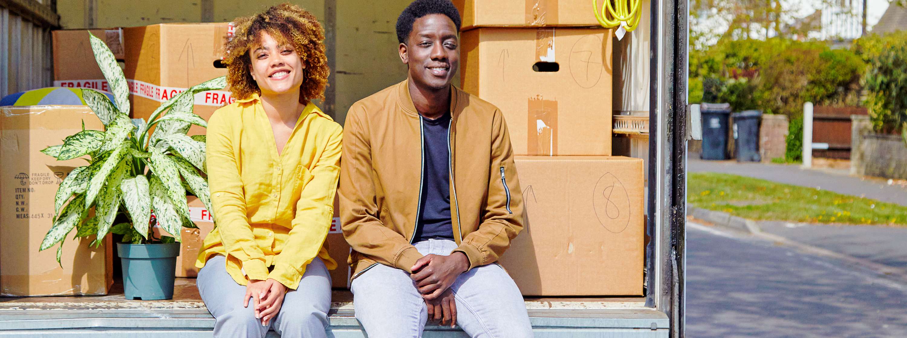 Young couple smiling and sitting in the back of an open moving van. It's a sunny day and the van is full of cardboard boxes.