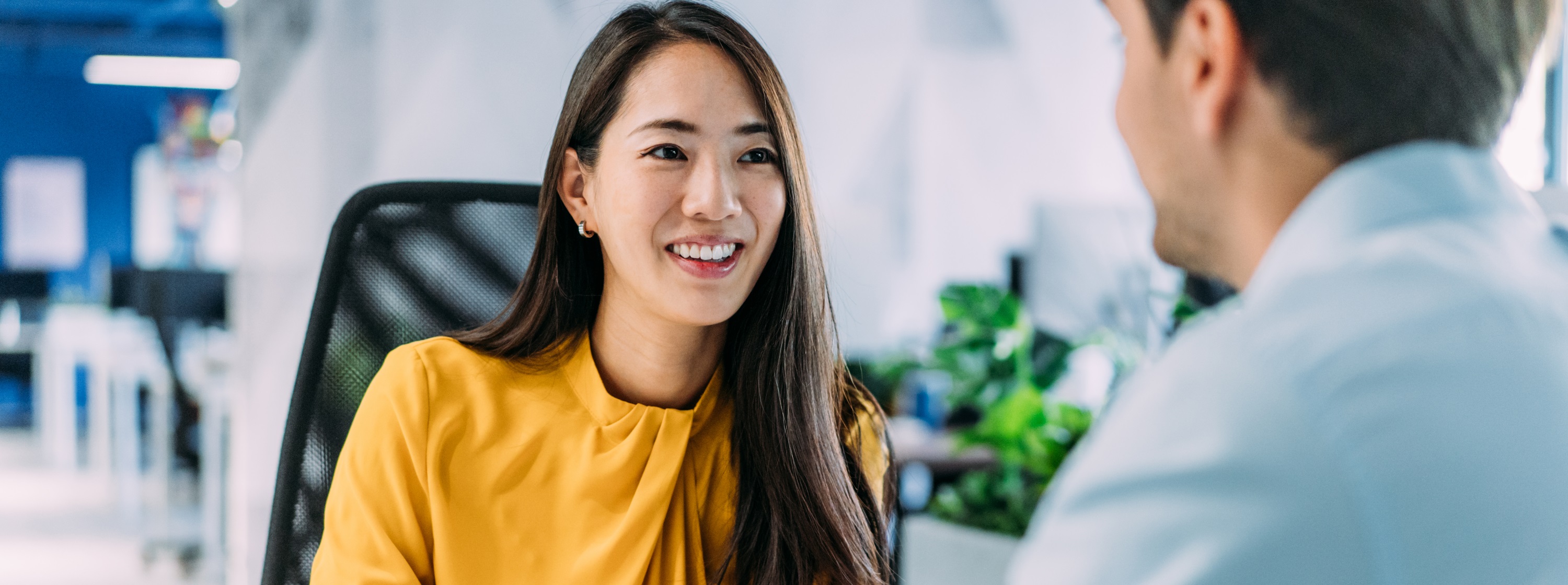 Smiling woman in a yellow blouse speaking with a colleague in an office setting