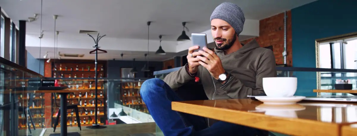 A young man with medium skin tone and a tidy beard and moustache, sits with one leg folded over the other. He holds his phone with both hands, smiling slightly as he types. He wears jeans, boots, and a beanie hat, sitting beside an oversized mug, in a bright coffee shop.