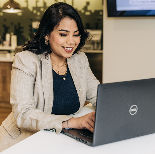 Woman using a laptop. She is smiling and she has dark hair and medium skin tone.