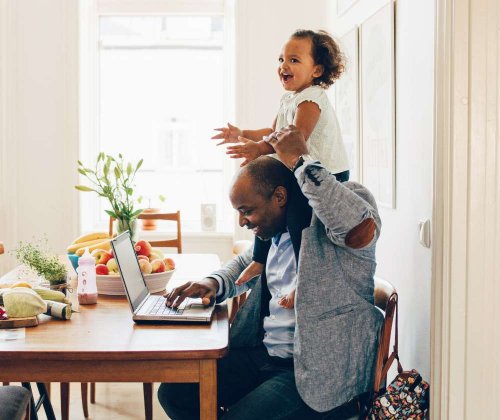 Smiling man sitting at his kitchen table with his laptop open. His daughter is sitting on his shoulders, laughing.