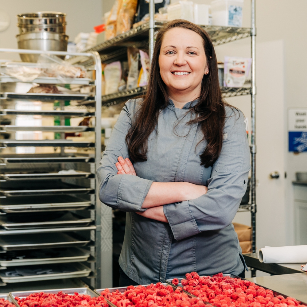 A light skinned woman at her place of business, a bakery. She is standing beside a tray of desserts.