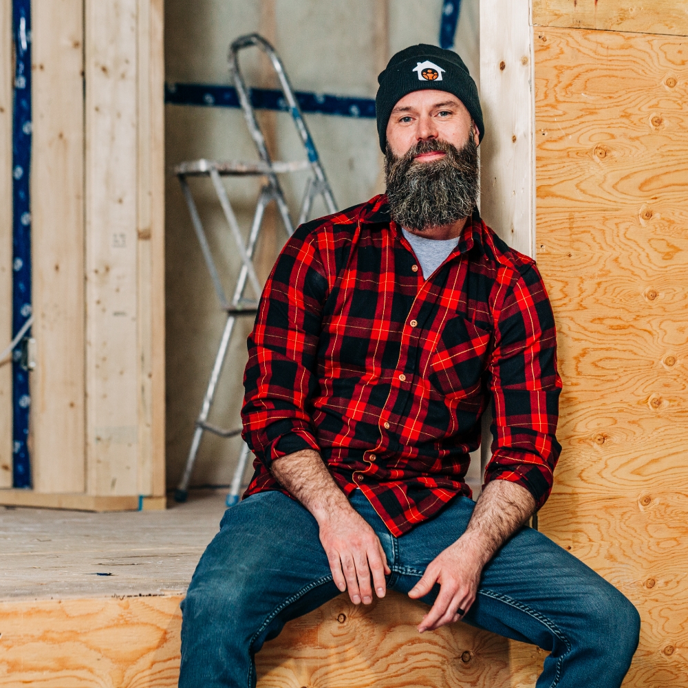 Light skinned man, a Staff Member from Community Builders, sits in a workshop. He’s wearing a toque and a plaid work shirt. 