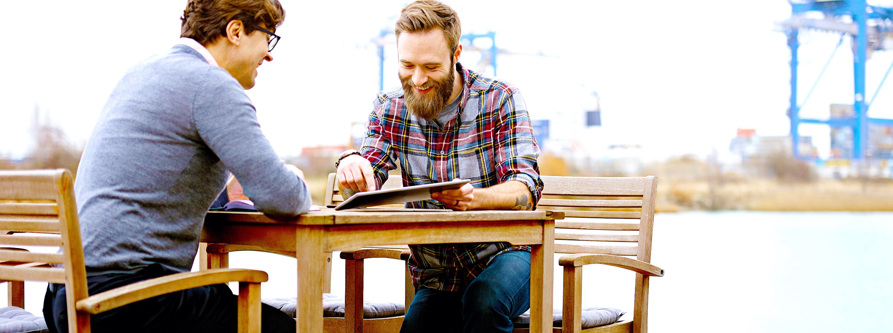 two males sitting at a table outside near an industrial site looking at a tablet