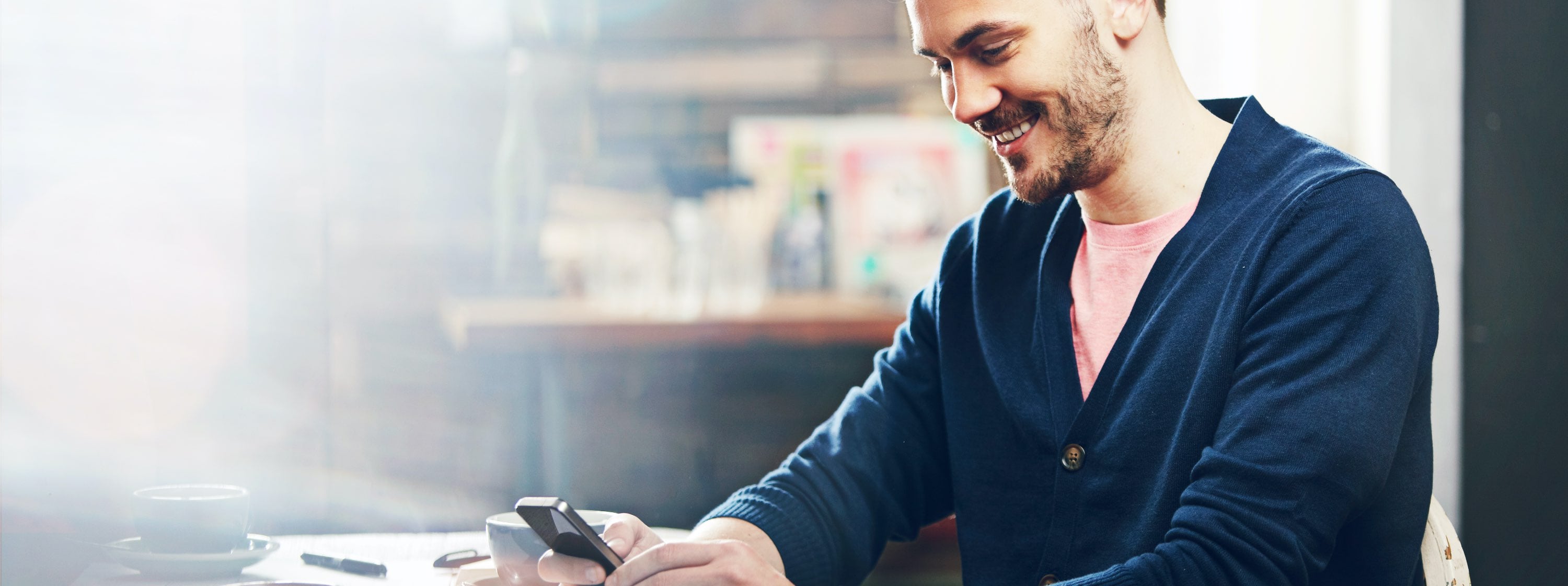 A young man with medium skin tone and a tidy beard, sits in a coffee shop. He holds his phone with both hands, smiling slightly as he types. 
