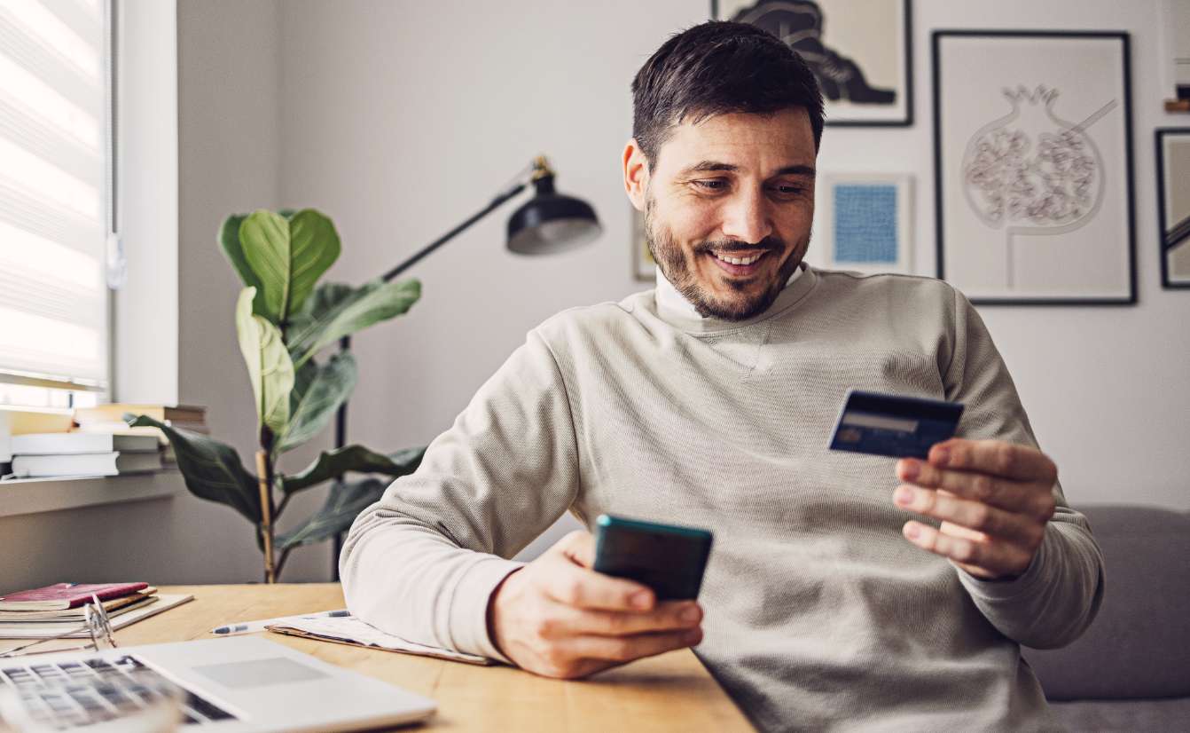 Smiling man at a desk holding a credit card and looking at his phone.