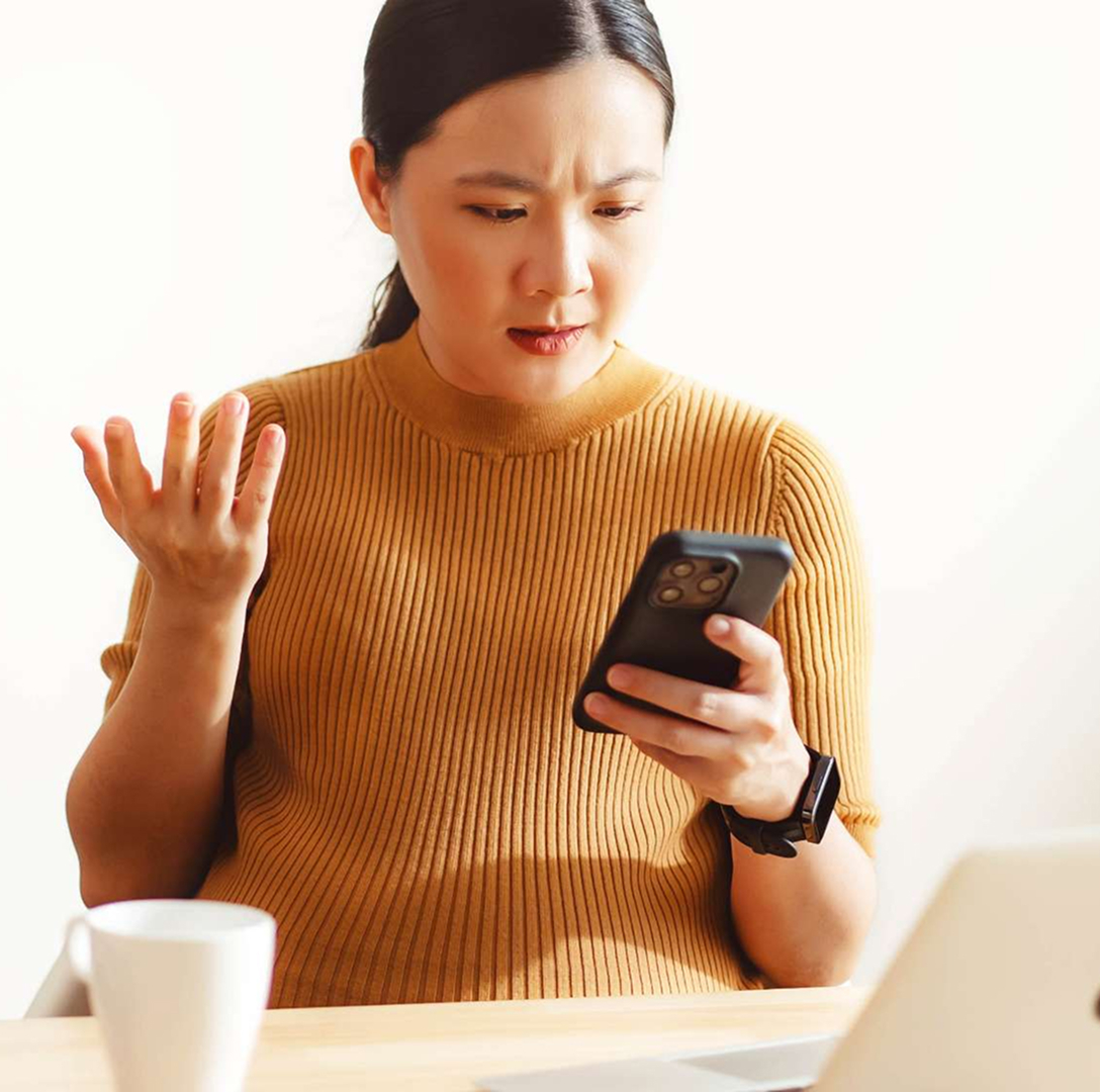 A young woman with medium skin tone and long dark hair sits at a desk, frowning. She is holding her phone in one hand and gesturing with the other.