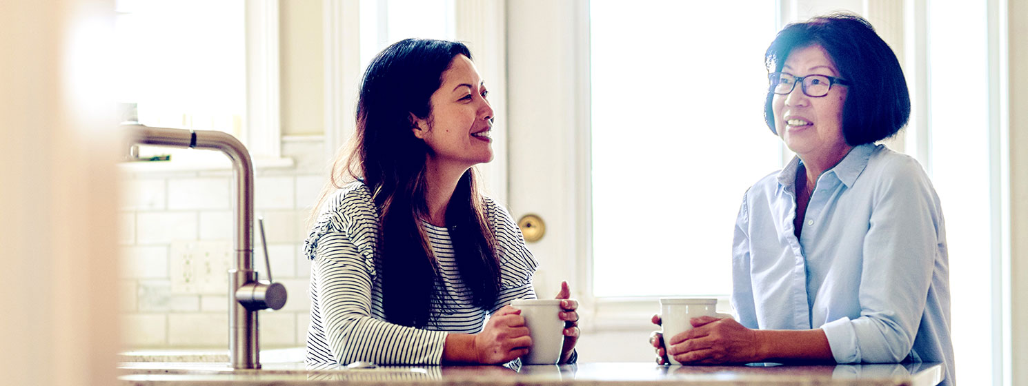 Two women sit in a bright kitchen, talking and drinking coffee.