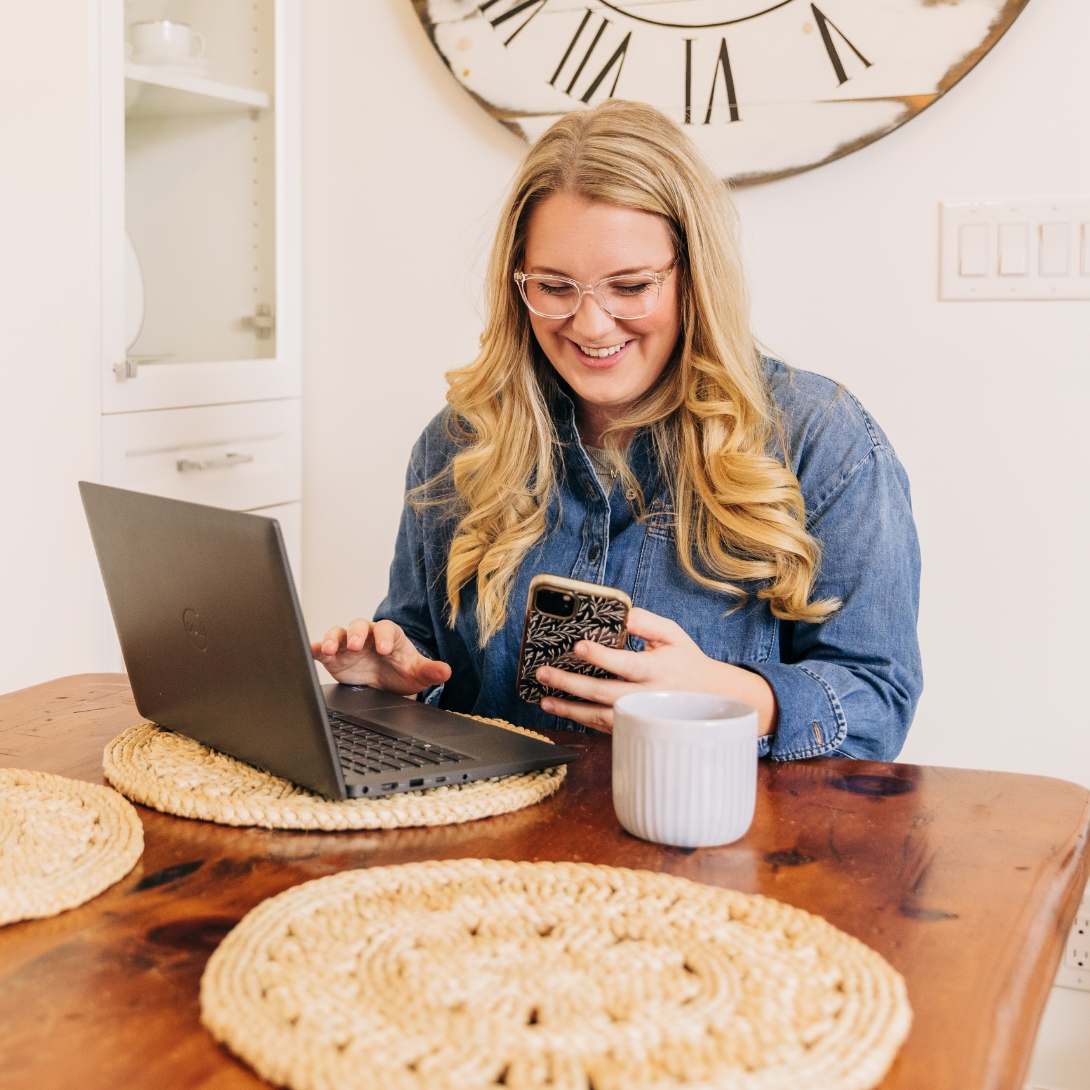 A smiling woman sits at a table using a laptop and a mobile phone.