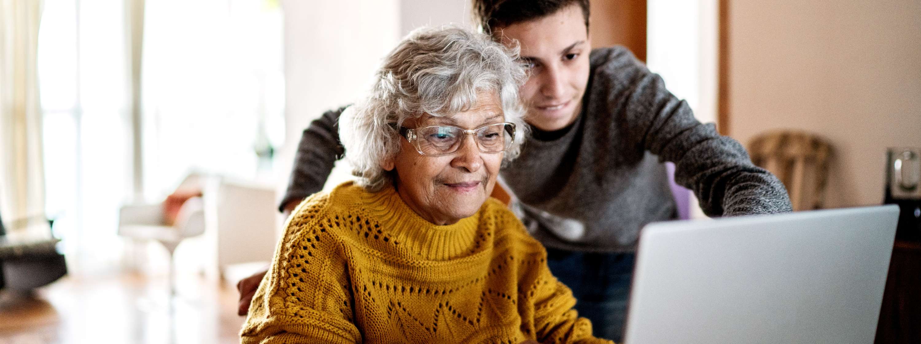 Older woman sits at a table with her laptop open. Her grandson is standing behind her, pointing at the laptop screen. 