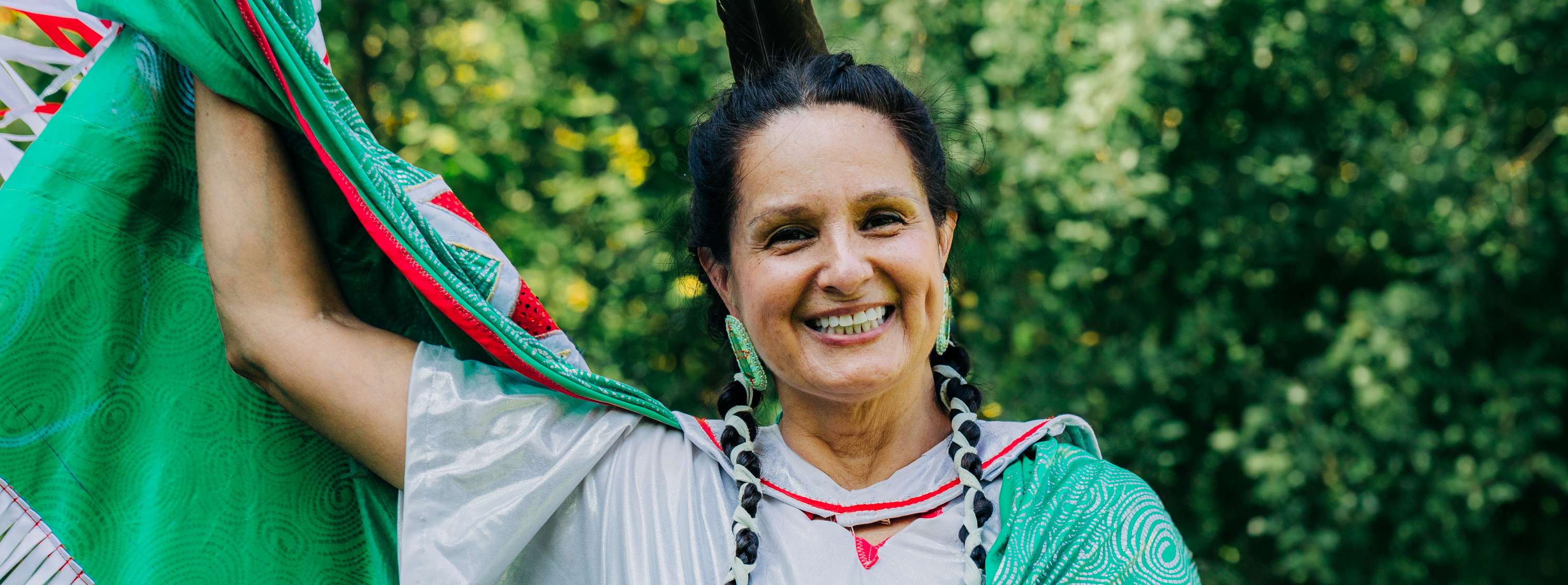Outside, surrounded by greenery, Cher Obediah smiles at the camera, wearing traditional indigenous regalia in green, red and white. With her hair in braids, she holds up one arm, sharing the colour and beauty of her regalia. 