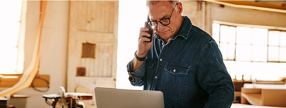 Older man in a workshop, talking on the phone and looking at a laptop