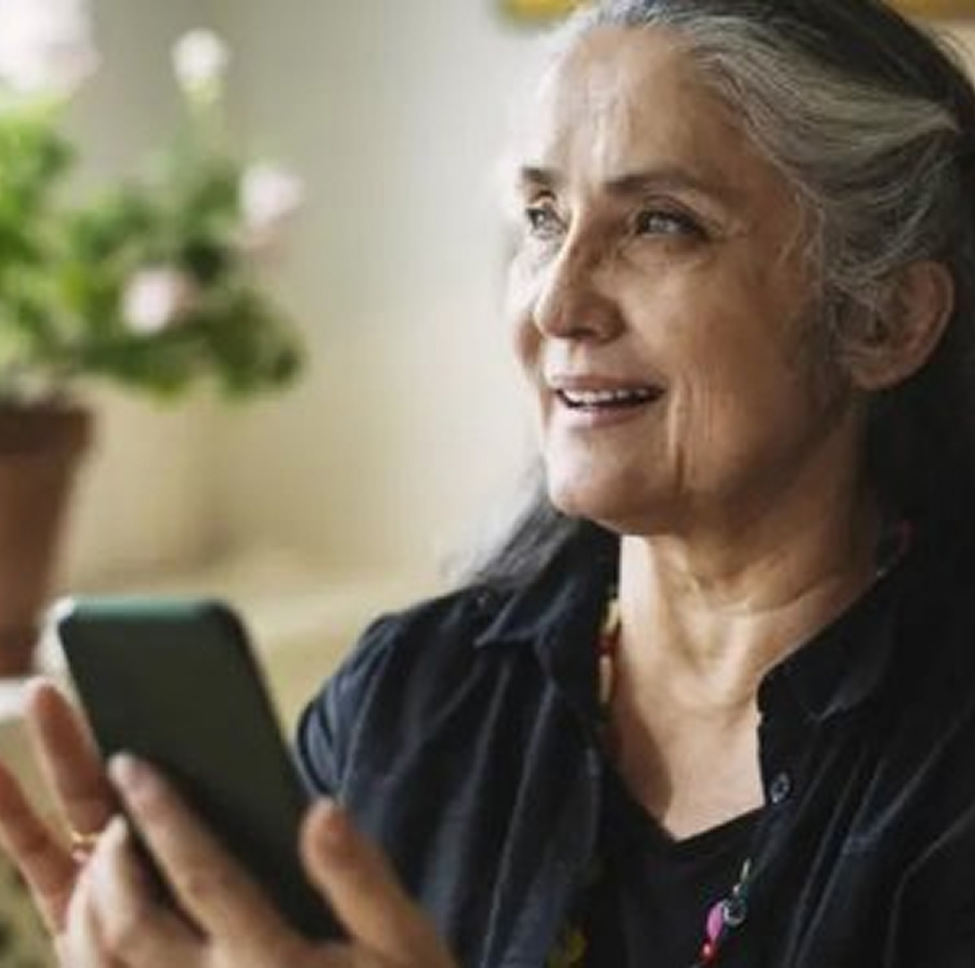 A mature woman with long, greying hair, holds her phone in front of her, smiling slightly as she speaks.
