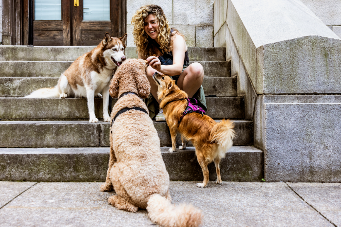 Young woman with fair skin and curly blonde hair sitting on a stoop, petting three dogs.