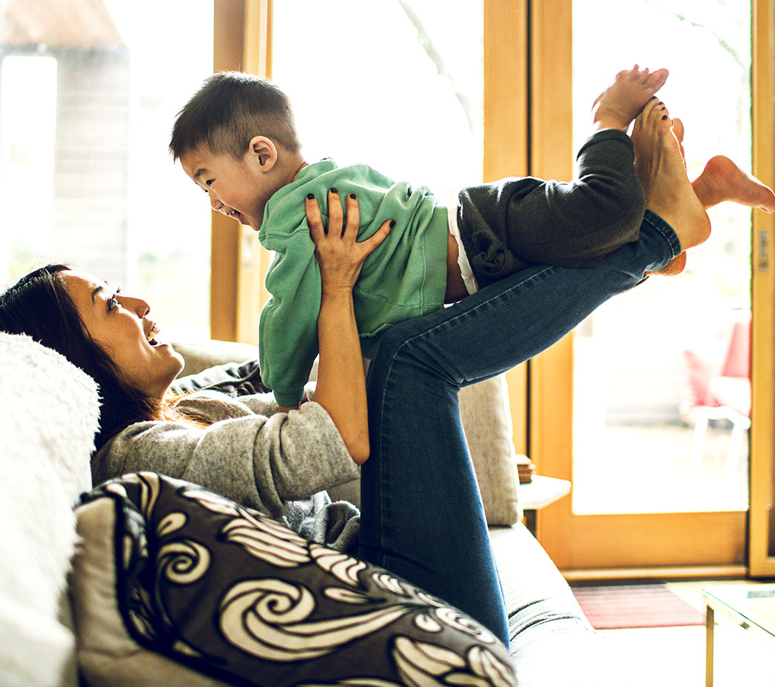 Mother plays with her young son on her lap, sitting on the sofa