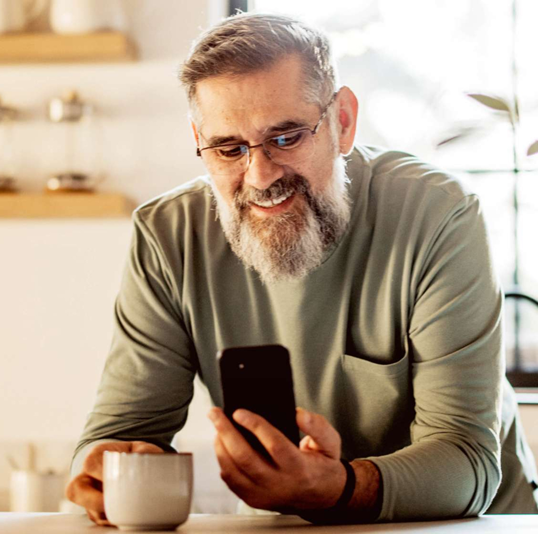 A middle-aged man with grey hair and long beard, sitting at a kitchen table holding a smartphone in one hand and a cup in the other, with natural light coming through the window behind him.