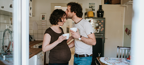man kissing pregnant wife's forehead, both holding coffee mugs in their kitchen
