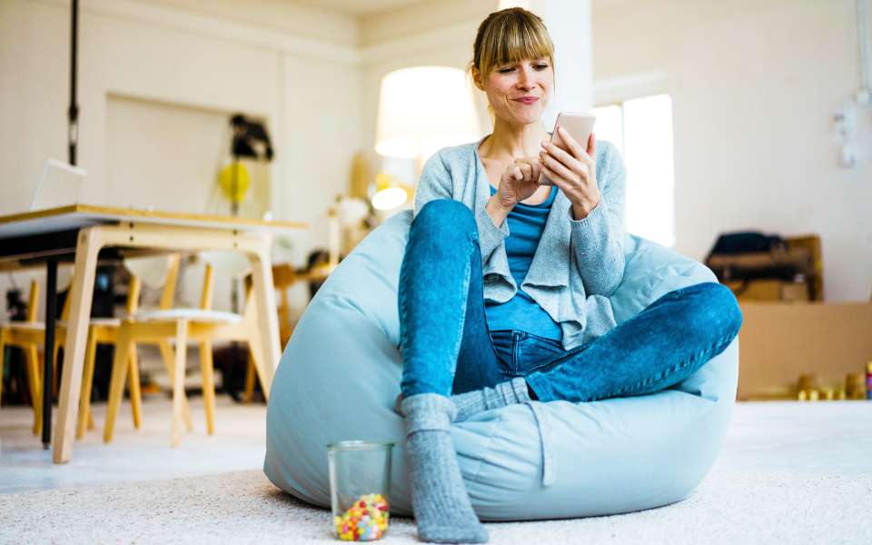 A woman lounges cross-legged on a bean bag chair at home, holding her mobile phone.