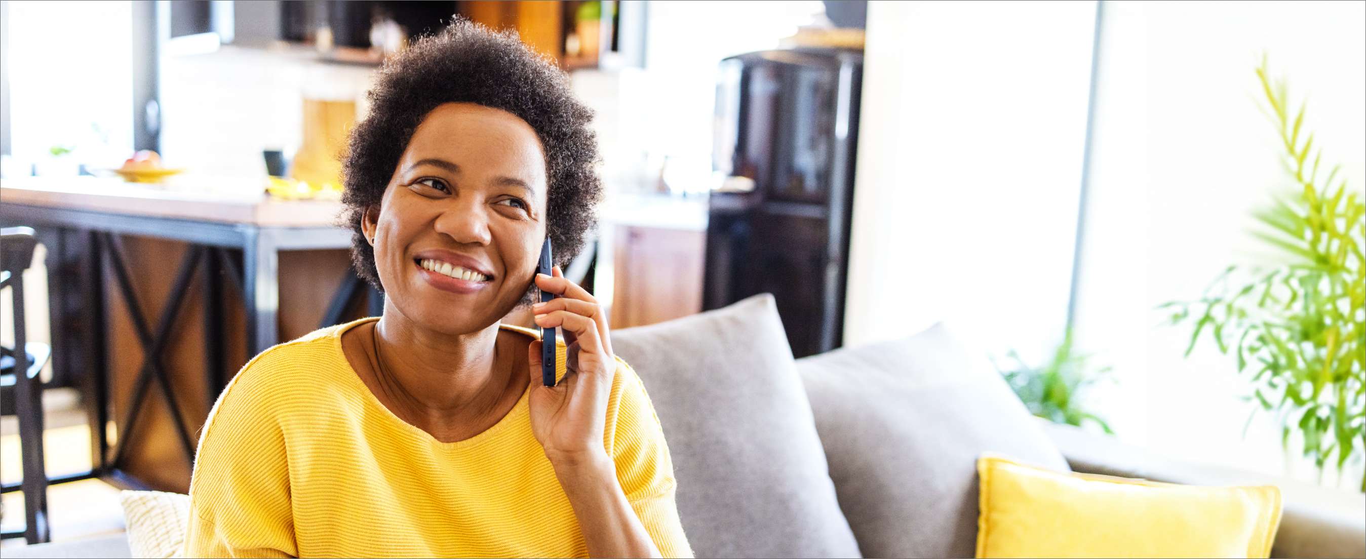 Mid adult black woman in yellow shirt sitting on the couch in the living room and talking on the phone, smiling.