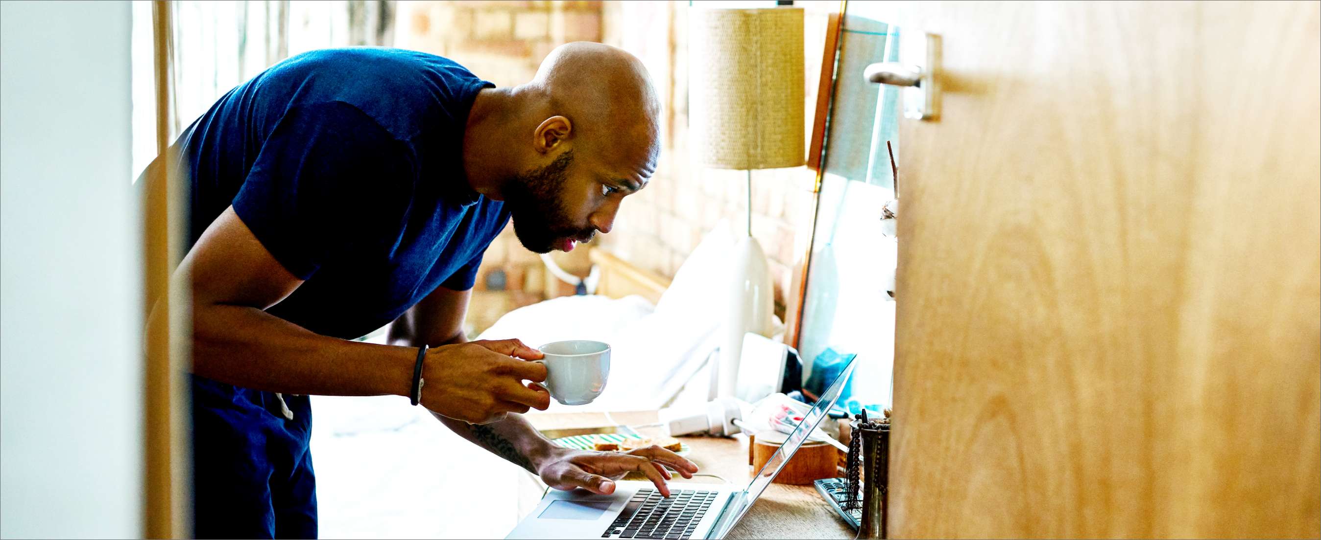 Mid adult black man using computer on dressing table in bedroom, holding mug, having breakfast whilst getting ready.