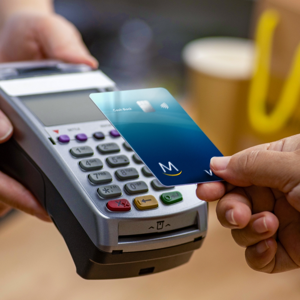 	 Close up of a hand holding a blue Meridian bank card. The card is placed next to a payment machine in order to tap and make a purchase. 