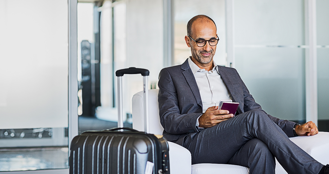 Man with suitcase, sitting comfortably in an airport lounge, looking at his phone.