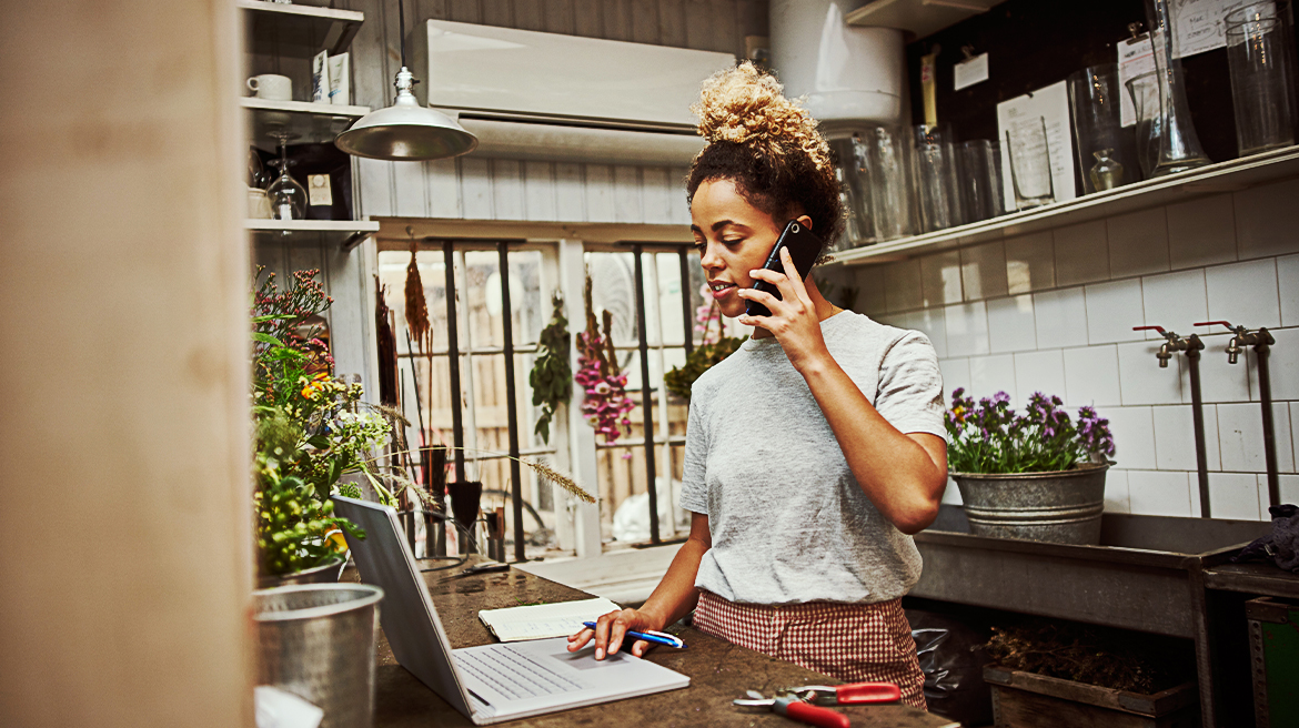 A woman in her kitchen talking on the mobile phone while working on her laptop