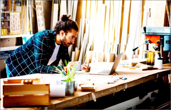 A young man with medium skin tone, dark hair pulled back, and a beard, looks intently at a laptop on his workbench in a sunny workshop.