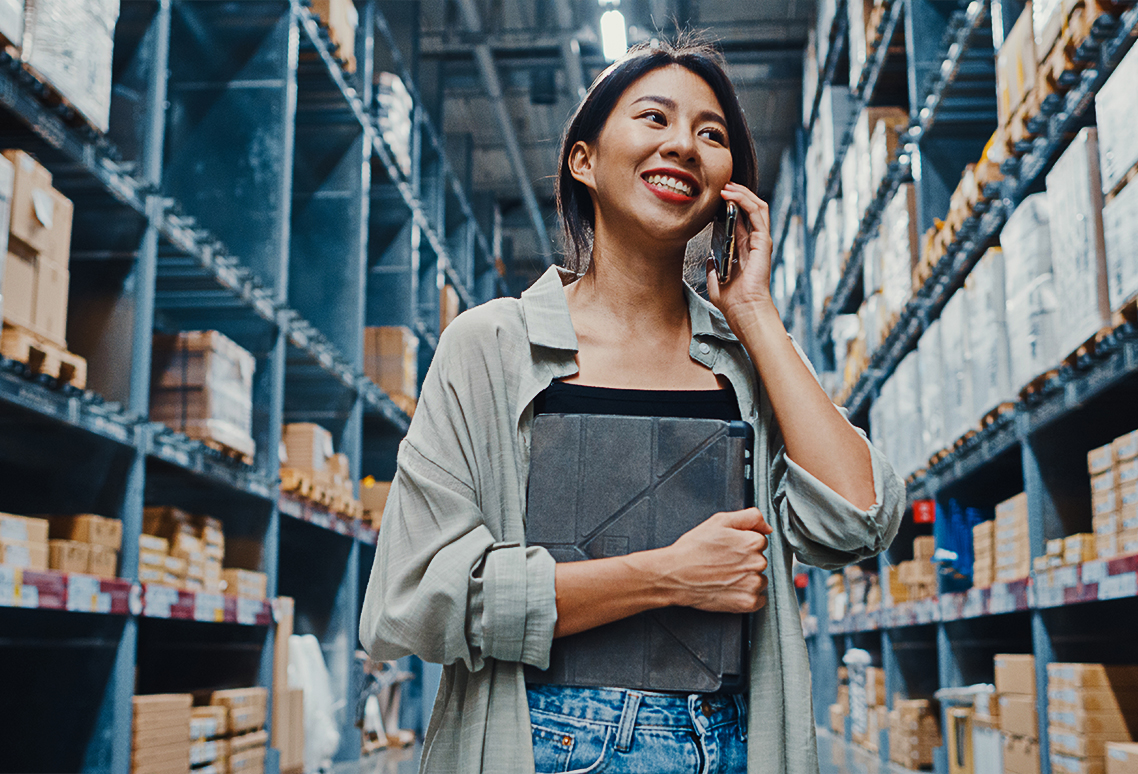Young woman at work in a warehouse, on the phone with clipboard.