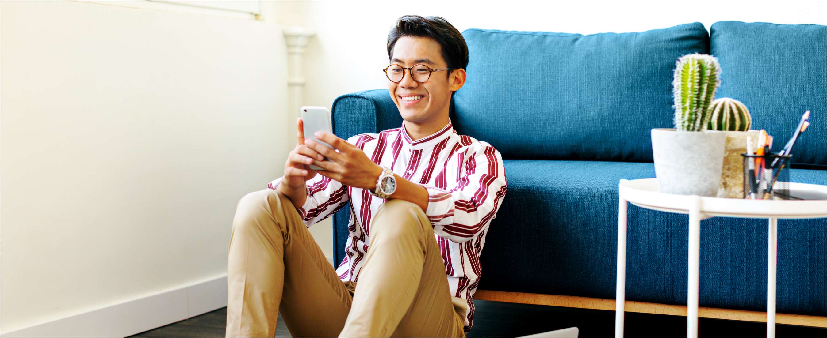 A young Asian man with glasses sits on the floor of his living room. His back is pressed up against a blue couch and he's smiling at his phone.