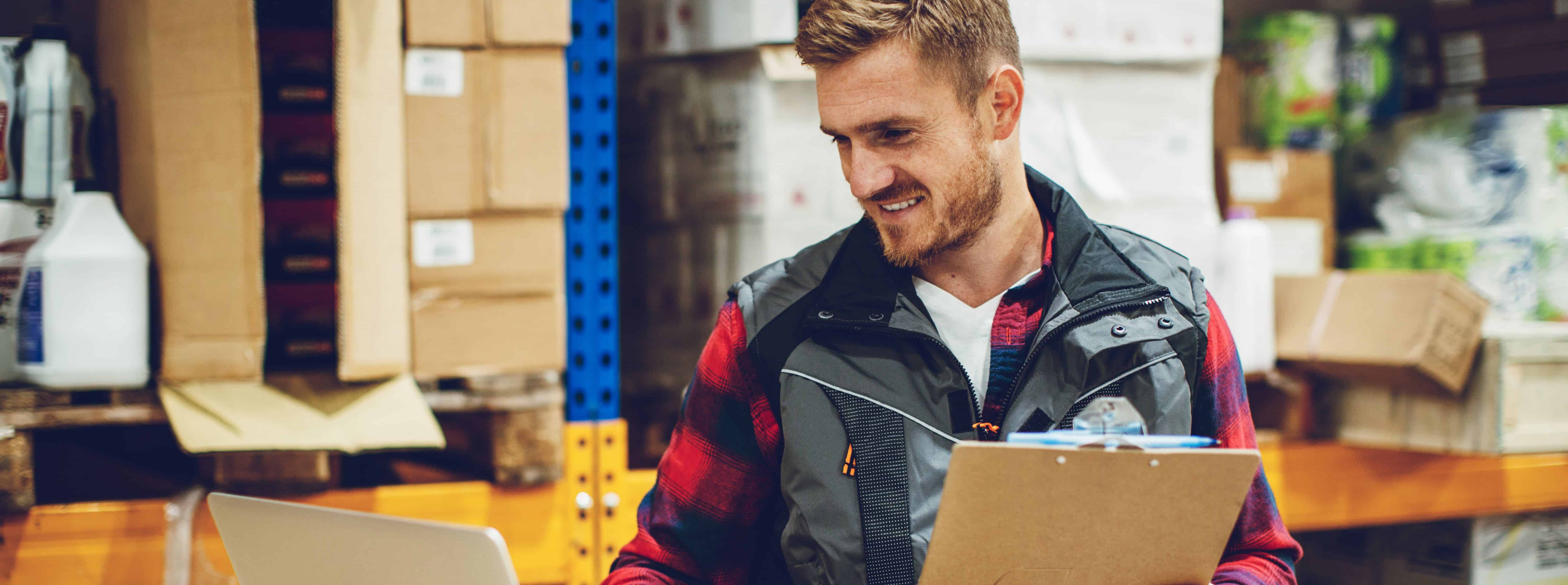 A smiling man with light skin and blonde hair holding a clipboard, using a laptop and working in a warehouse.