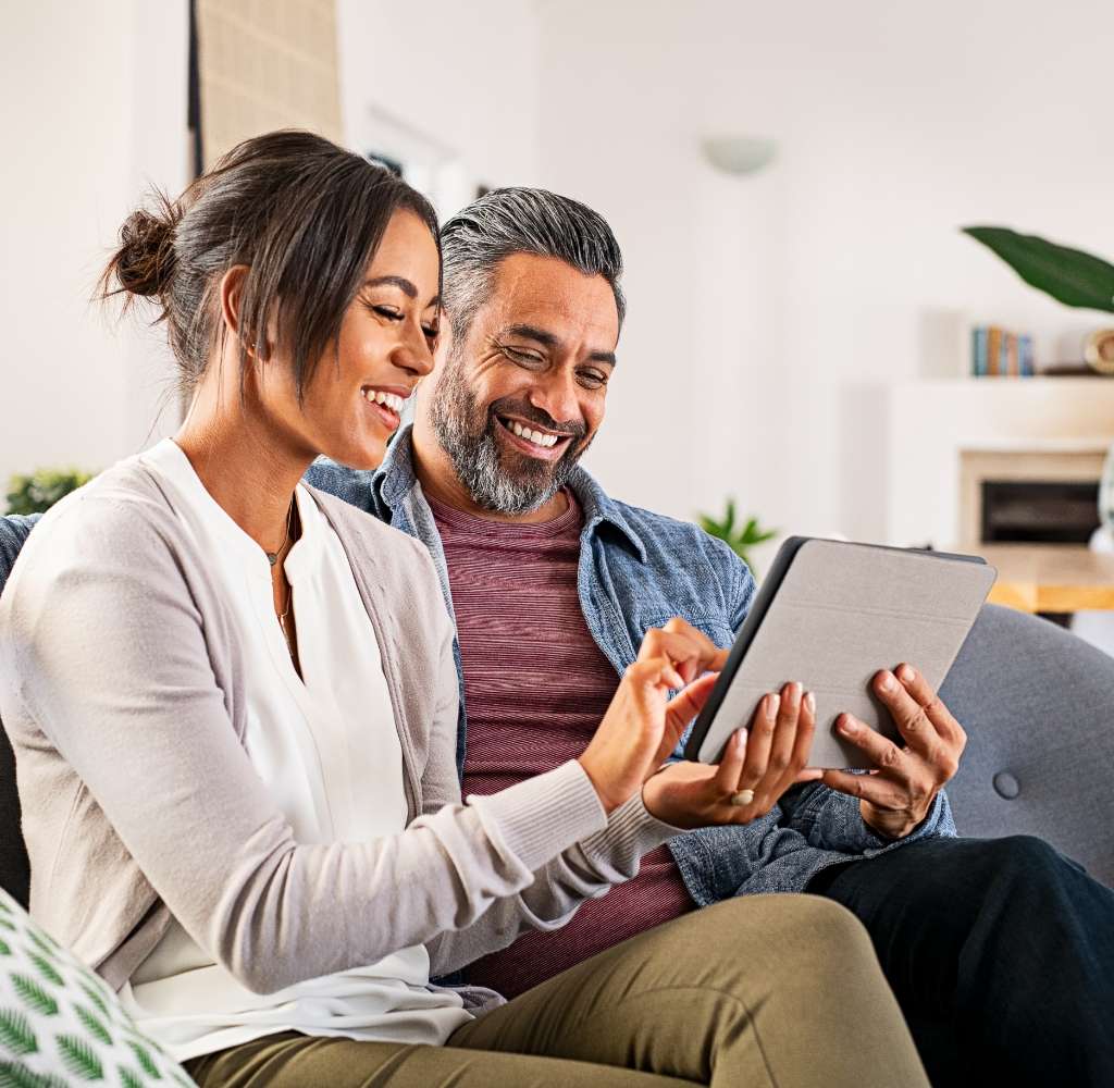 A couple sit together on the sofa at home. They’re smiling and using an tablet.