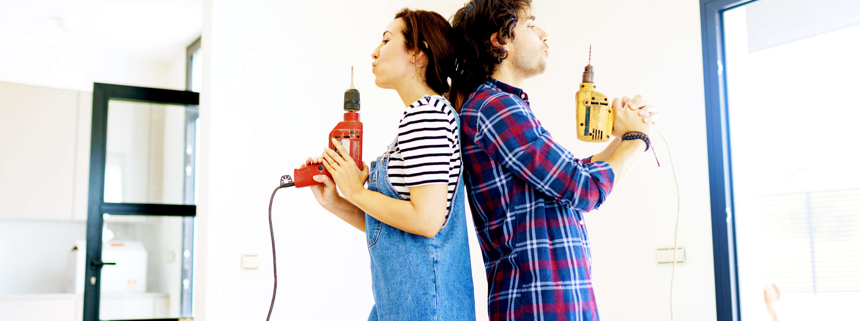 A couple about to start a renovation project at home. They stand back to back, each holding a drill like a smoking gun.