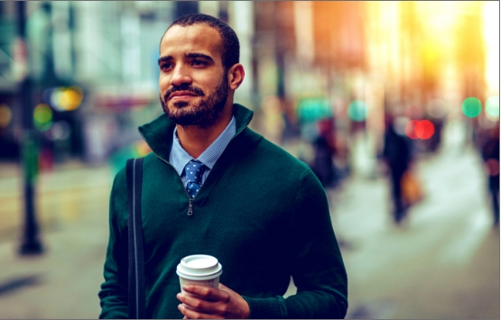 A young man with medium skin tone, short dark hair and a beard walks down a city street with a cup of coffee.