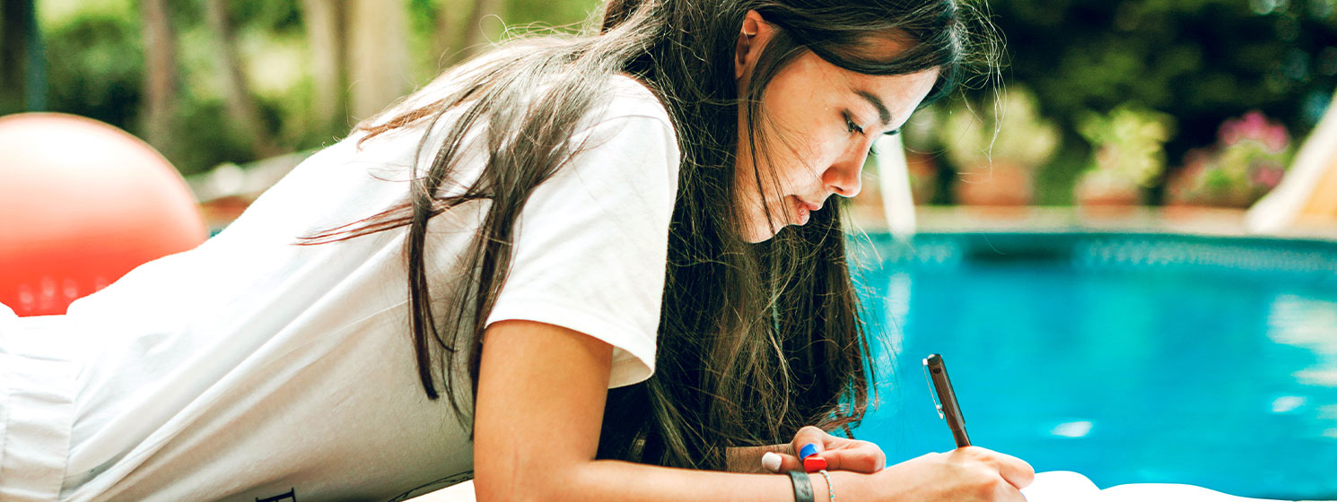 Teenage girl writing in a notebook. She is laying outside by a pool