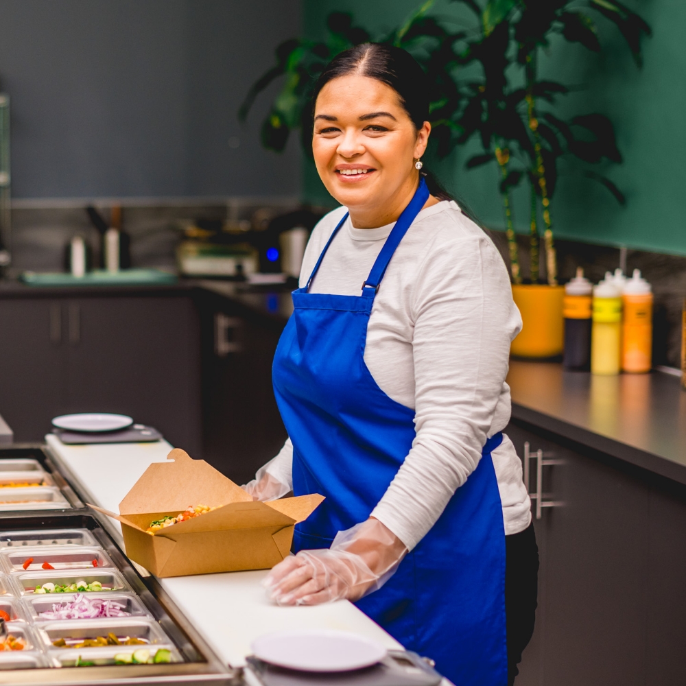 Staff member at Urban Cravings is behind the counter, wearing a blue apron, putting a take out box together. 