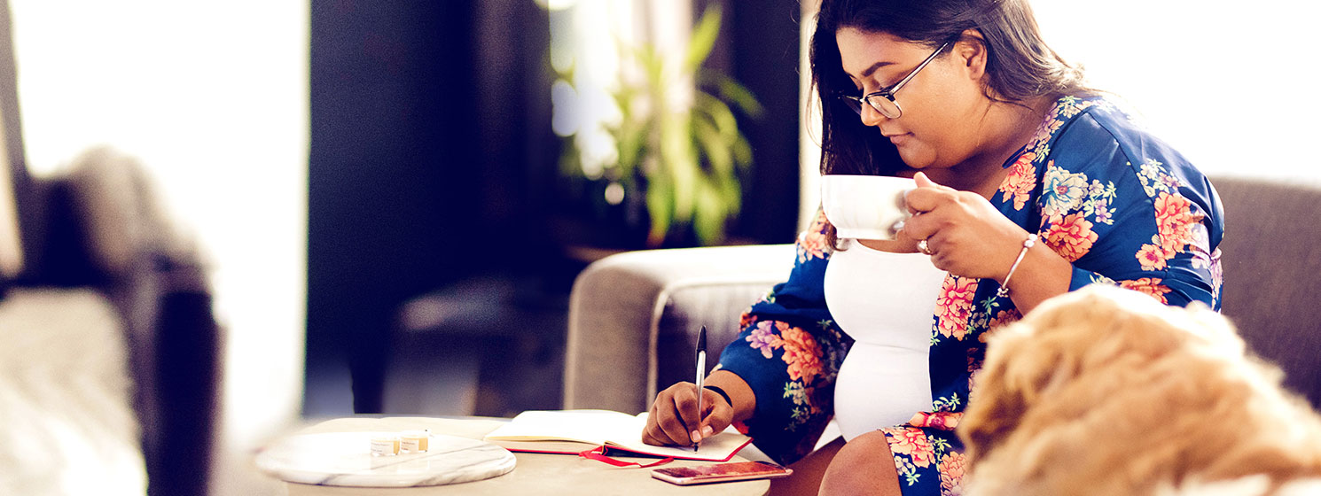 Woman writing in her notebook while sitting in living room with a cup of coffee