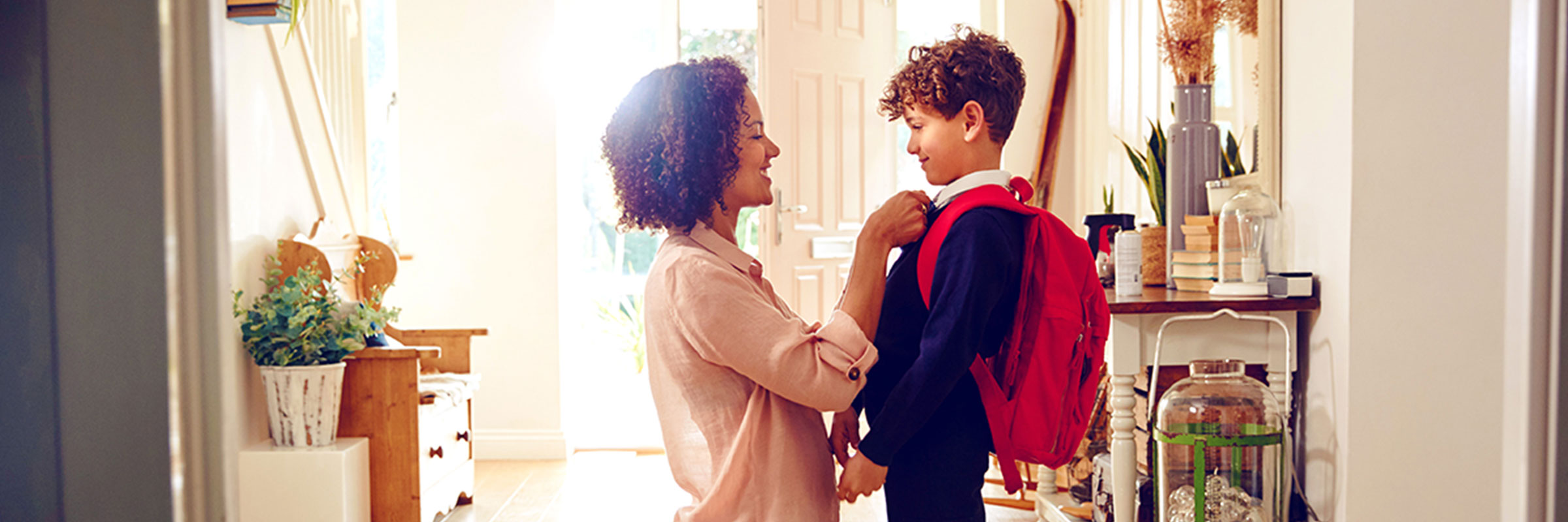Mother and young son standing in the doorway of their home, getting ready for school. She is fixing his uniform and he is wearing a red backpack.
