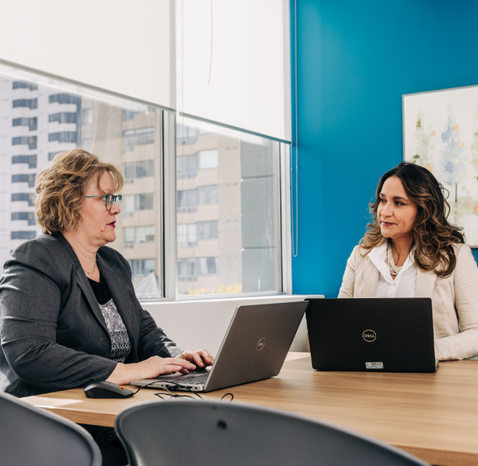 Two women chat while working on laptops in an office.