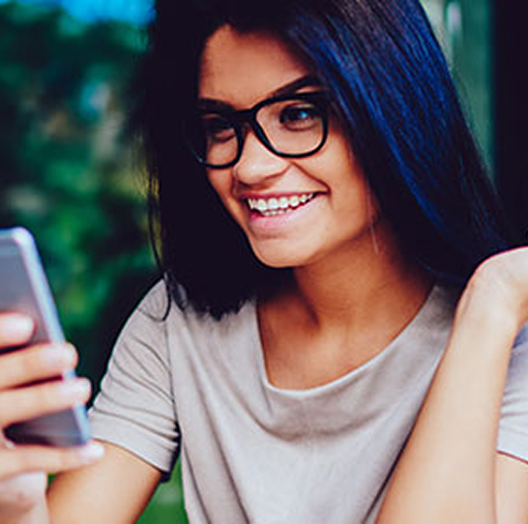 Young woman with dark hair, medium skin tone, and glasses smiles at her phone while sitting in an outdoor cafe.